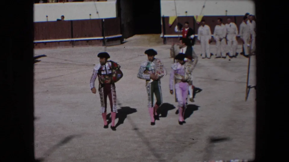 Bullfighters in ornate suits and pink stockings proceed across the sunlit Barcelona bullring, with a line of attendants standing near the arena wall.