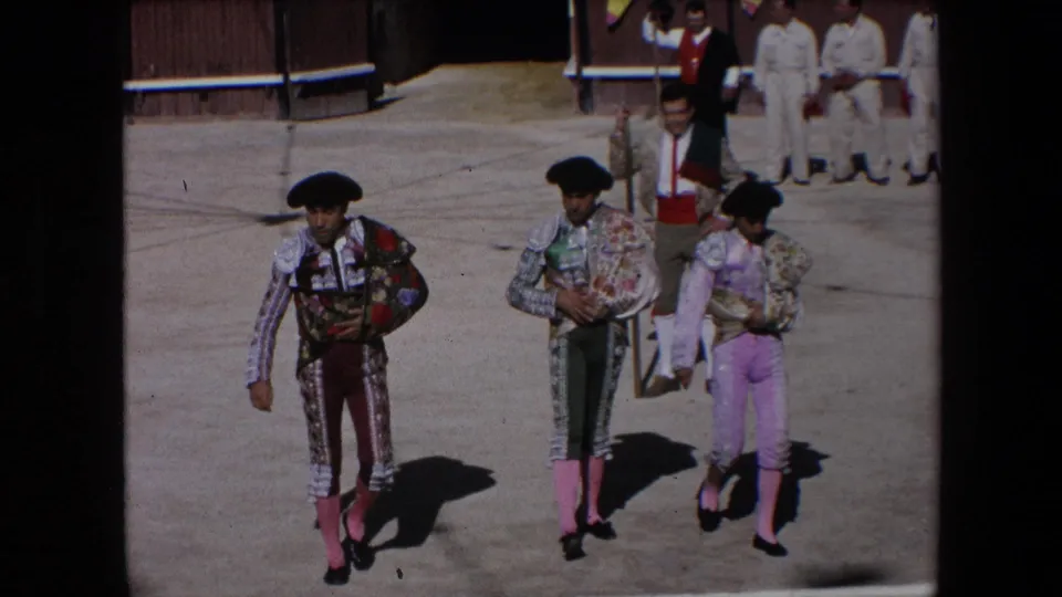 In Barcelona, Spain in 1971, three matadors in ornate trajes de luces and black monteras walk across a bright bullfighting arena floor with attendants in the background.