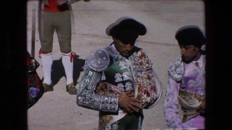 A close spectator-angle view shows two matadors in ornate suits and black monteras walking near the arena edge, with part of another costumed figure at the left.