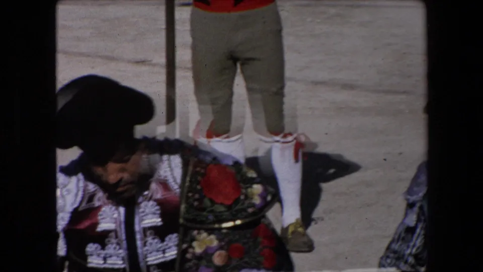 1971 Barcelona frame showing a matador in ornate bullfighting costume and black hat, partly cropped beside another figure's white socks with red trim on the arena floor.