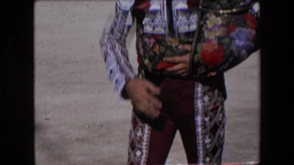 Close 1971 Barcelona detail of a matador's ornate bullfighting jacket with fringe, maroon trousers, and hand positioned at the waist in bright arena light.