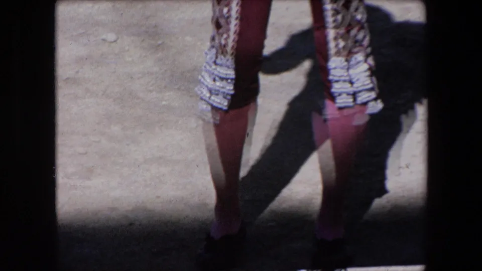 1971 Barcelona frame focused on the lower half of a matador's costume, with maroon trousers, embroidered jacket panels, and strong shadow across the arena surface.