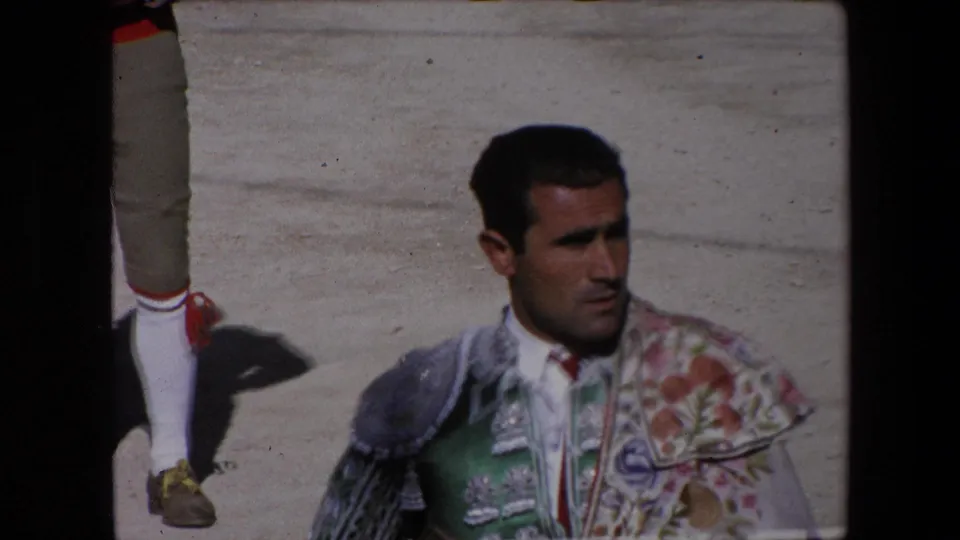 Adult torero in green and floral embroidered bullfighting attire stands in bright sun on arena sand in 1971 Barcelona footage, with another participant partly visible at the edge.
