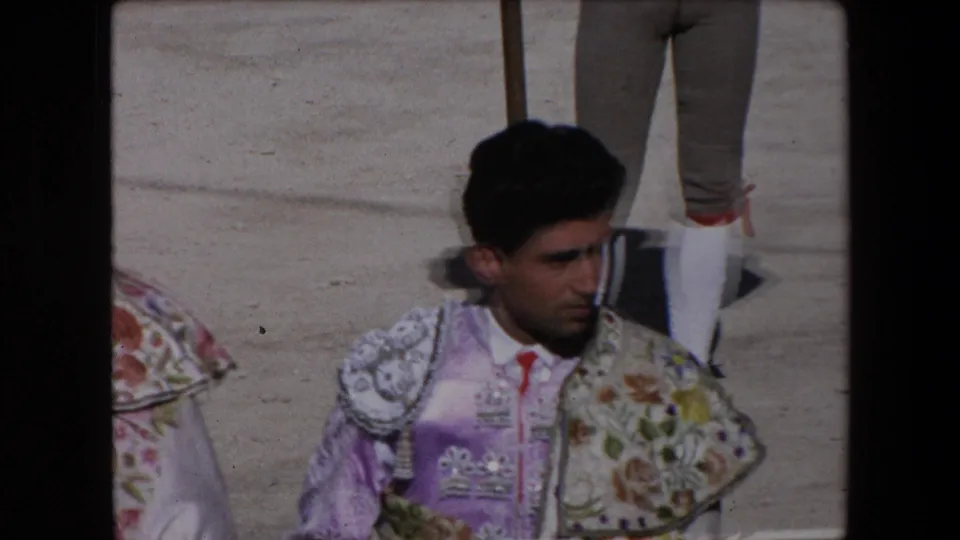 Adult torero in purple and floral bullfighting attire stands on the arena sand, partly framed by another torero and nearby legs in the 1971 Barcelona footage.