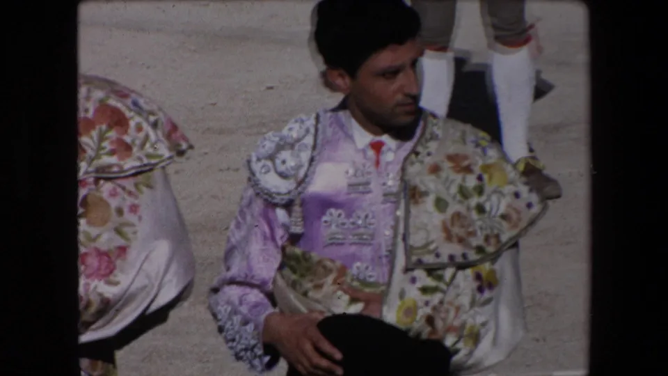 Adult torero in purple and floral embroidered attire holds a black montera-style hat while standing in the sunlit Barcelona bullring footage from 1971.