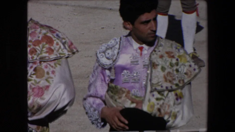 In Barcelona 1971 8mm footage, a matador in a purple and floral embroidered suit stands in partial close-up beside the sandy arena area.