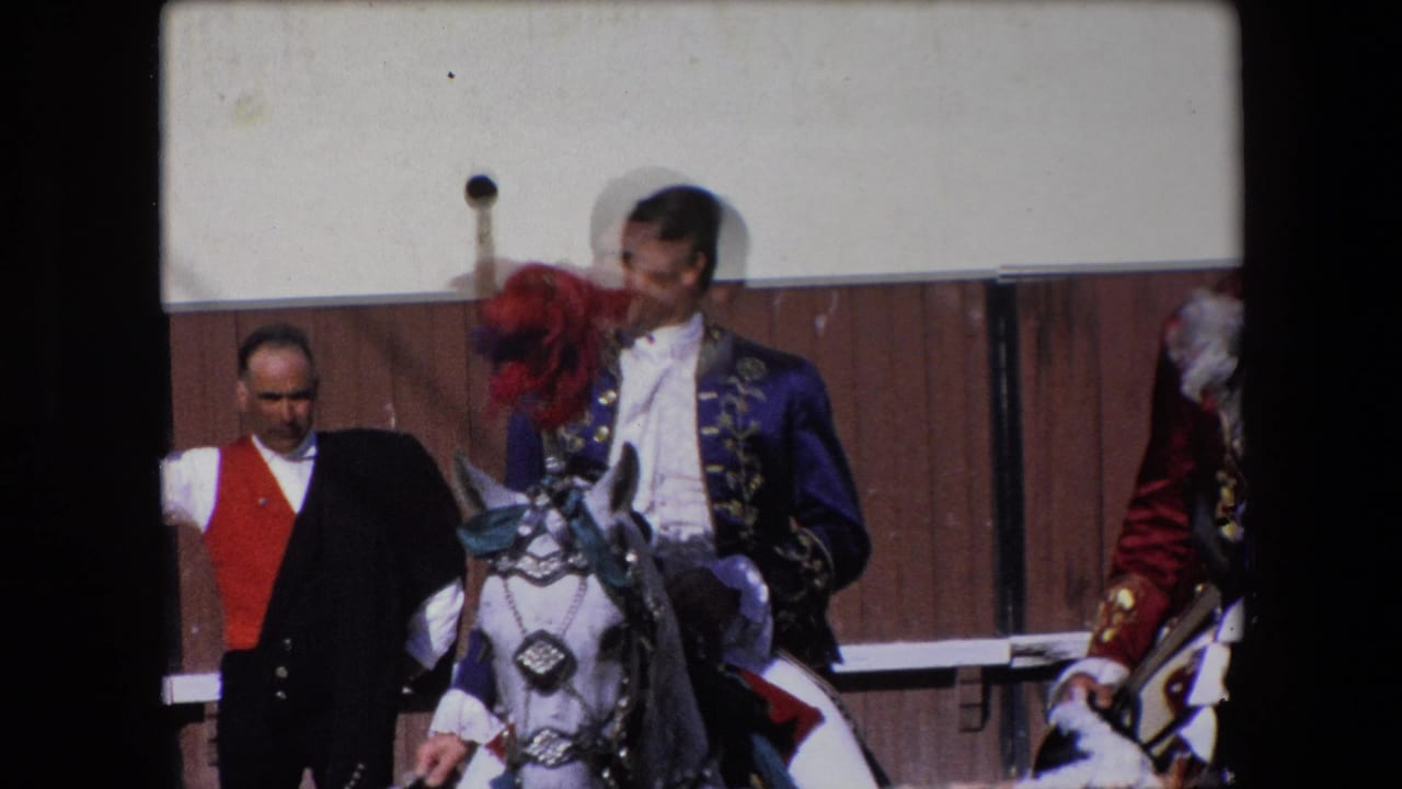 A 1971 Barcelona home-movie frame shows a rider in an ornate blue jacket on a white horse with a red plume, with an attendant standing near a wooden arena fence.