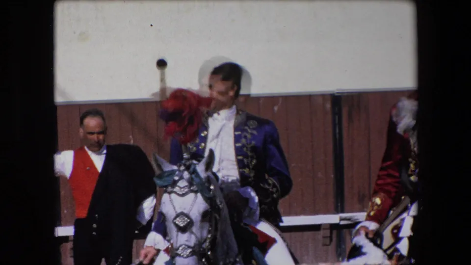 A 1971 Barcelona home-movie frame shows a rider in an ornate blue jacket on a white horse with a red plume, with an attendant standing near a wooden arena fence.