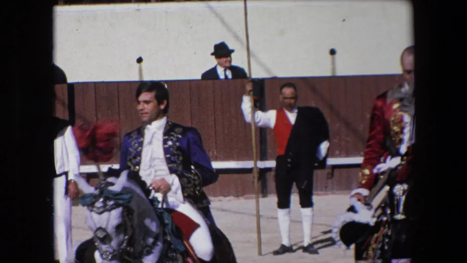 A costumed rider on a white horse appears in the Barcelona arena footage, with attendants, a pole, and an observer above the wooden fence visible in the background.