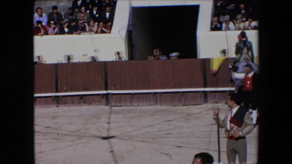 1971 Barcelona 8mm frame showing a mounted rider in green-and-gold dress near a bullring entrance, with a man holding a pole in the foreground and spectators seated above the wooden barrier.