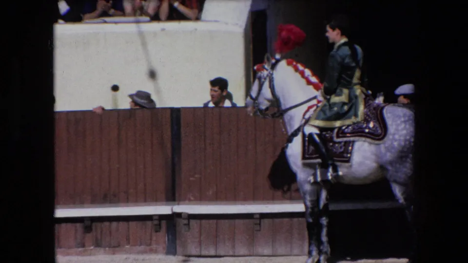 1971 Barcelona 8mm frame showing a rider in a dark green uniform on a decorated dapple-gray horse with a red plume, moving past the bullring barrier as nearby spectators look on.