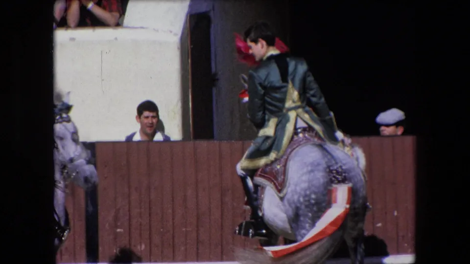 1971 Barcelona 8mm frame showing the rear view of a rider in a green-and-gold uniform on a decorated dapple-gray horse beside the bullring barrier, with several faces visible behind the rail.