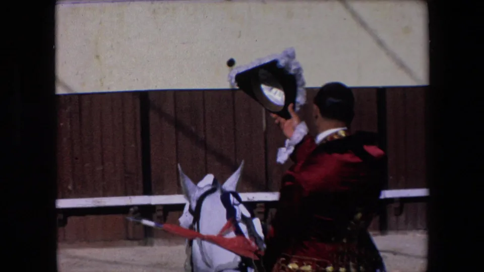In Barcelona in 1971, a bullfighting participant in a red and gold jacket raises a black and white hat in salute beside a white horse near the arena barrier.