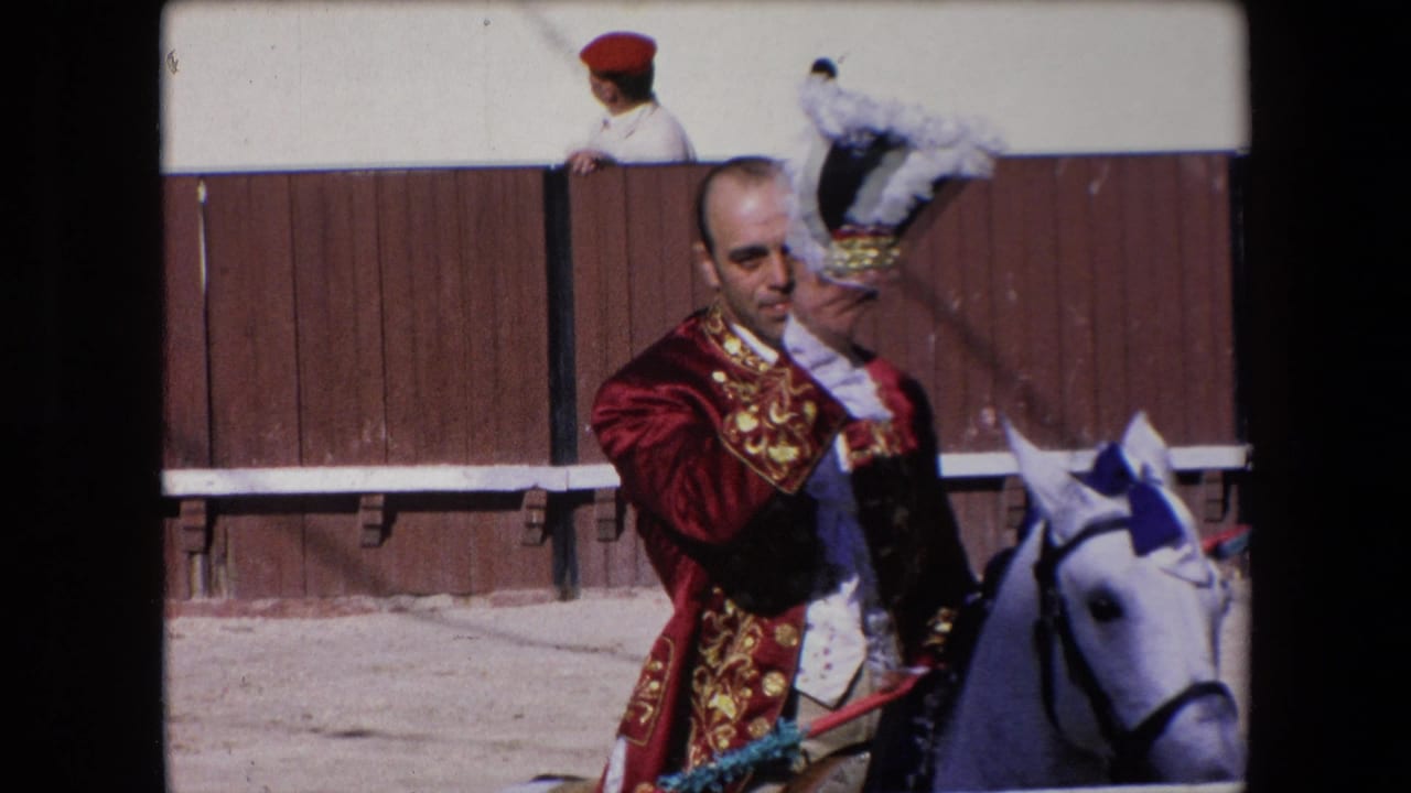 In Barcelona, Spain in 1971, a matador in a red and gold embroidered jacket rides a white horse and lifts a black-and-white feathered hat near a sunlit wooden bullring barrier.