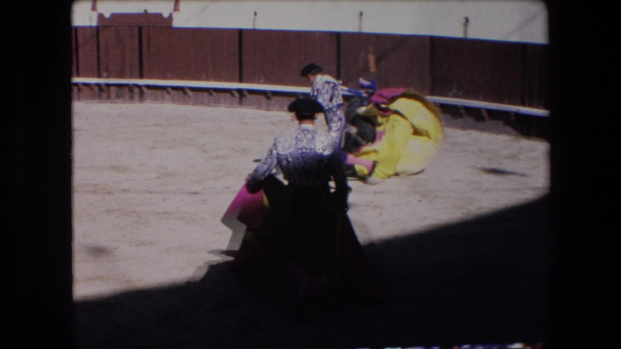 Two matadors in patterned suits stand with capes near a bull and bright yellow cloth during the Barcelona bullfight, seen from a shaded spectator angle.