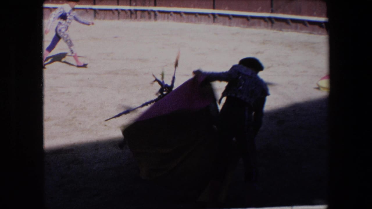 In 1971 Barcelona bullfight footage, a matador in ornate dress stands partly in shadow with a red cape as a bull approaches in the sunlit ring.