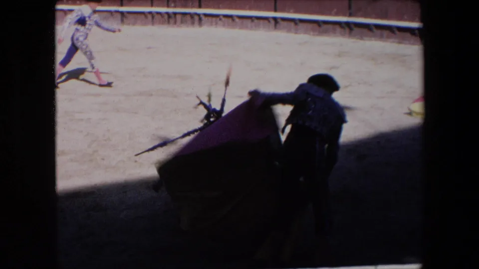 In 1971 Barcelona bullfight footage, a matador in ornate dress stands partly in shadow with a red cape as a bull approaches in the sunlit ring.