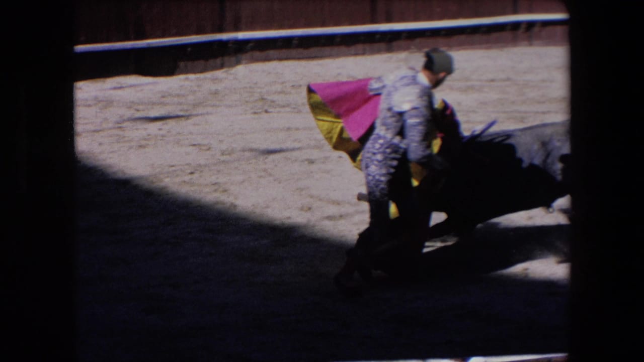 A matador leans near the dark bull while the red and yellow cape lifts in strong sun and shadow during the Barcelona bullfighting sequence.