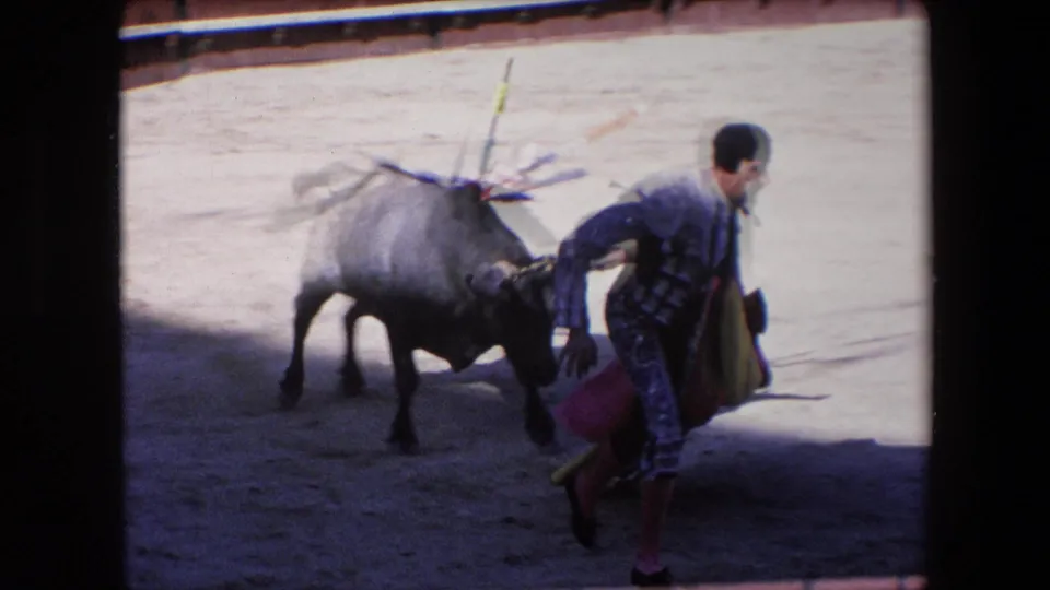 In the supplied 1971 Barcelona footage, a matador in ornate costume holds a cape while facing a dark bull on sandy arena ground.