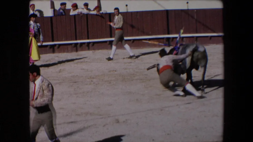 Barcelona, Spain, 1971 8mm frame showing a grey bull moving away while a costumed participant is low to the ground near the bull's hind legs.