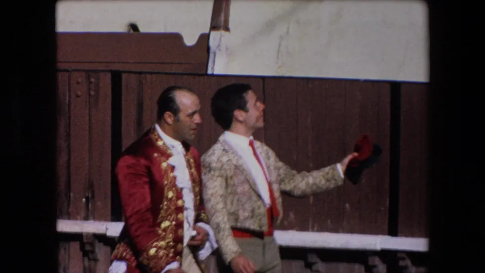 Two bullfighters in ornate jackets stand beside the wooden barrier in the 1971 Barcelona arena, with one holding a small red cap or cloth.