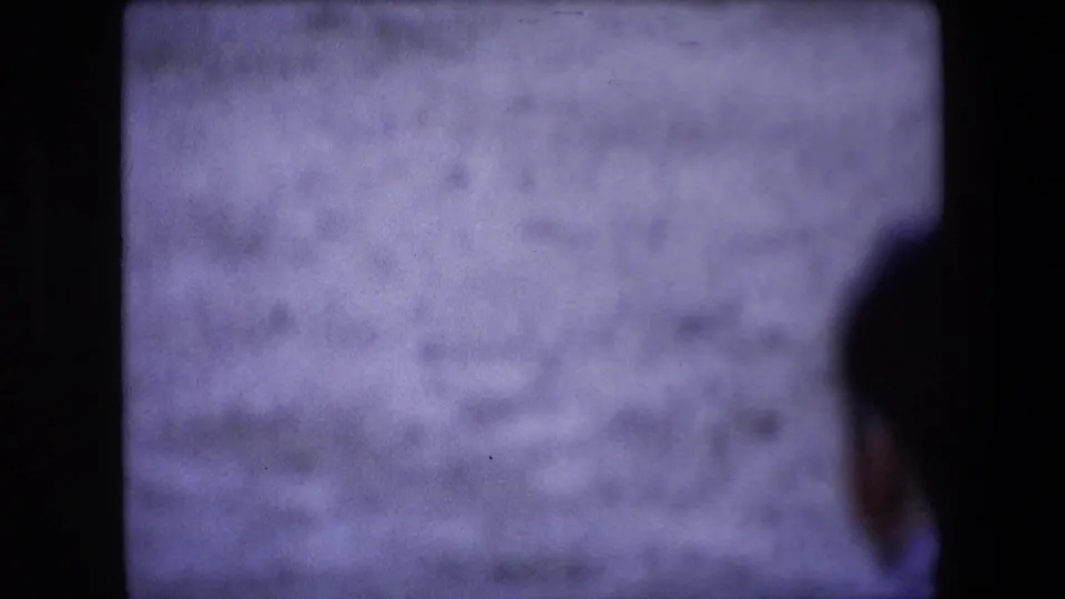 Soft-focus 1971 Barcelona home-movie frame showing a partially seen spectator or passerby against a pale arena-like wall, divided by dark film-frame borders.