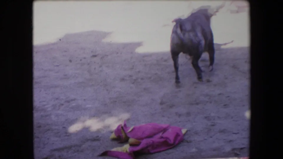 A black bull moves away across the pale sandy arena while a pink cape cloth lies in the foreground, framed by soft, grainy 8mm exposure.