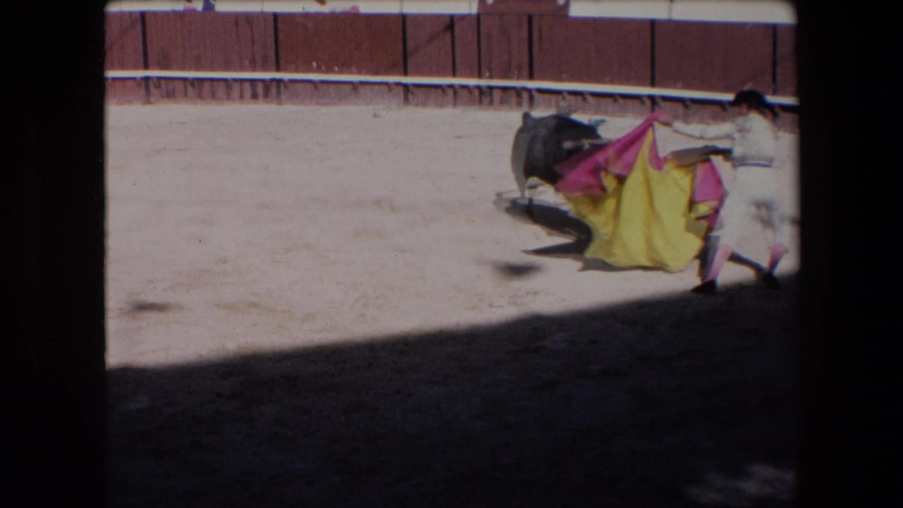 In the supplied 1971 Barcelona bullfight footage, a matador in a light outfit braces with a red and yellow cape as a black bull approaches across the sandy arena.