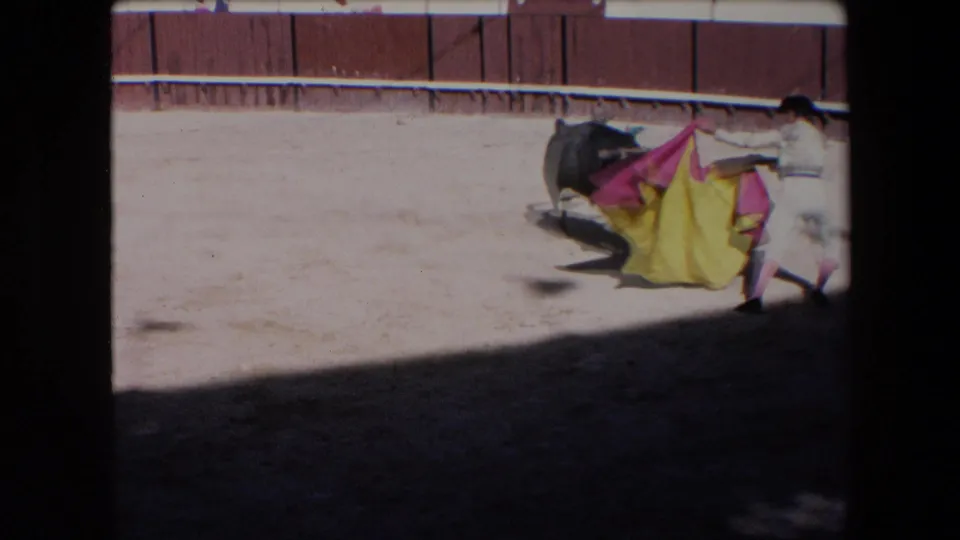 In the supplied 1971 Barcelona bullfight footage, a matador in a light outfit braces with a red and yellow cape as a black bull approaches across the sandy arena.