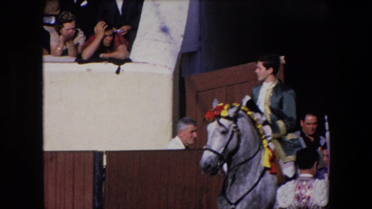 In 1971 Barcelona, a male rider in a dark suit with gold trim sits on a grey horse decorated with red and yellow ribbons near wooden arena panels and raised spectators.
