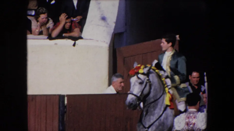 In 1971 Barcelona, a male rider in a dark suit with gold trim sits on a grey horse decorated with red and yellow ribbons near wooden arena panels and raised spectators.
