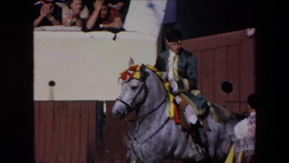 The rider appears to steady or guide the decorated grey horse as it moves along the wooden pen in the 1971 Barcelona home-movie footage, while spectators watch from the raised edge.