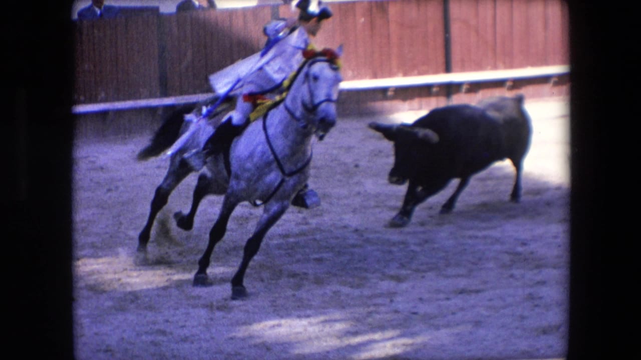 In Barcelona, Spain in 1971, a mounted rider on a grey horse leans through a turn while a black bull charges across the sandy bullring nearby.