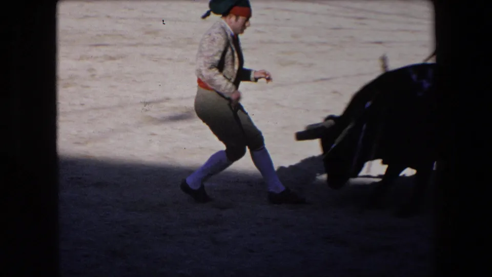 A bullfighter in cap, sash, and high socks steps near a charging black bull in the Barcelona arena, with the action partly shaded and blurred by motion.