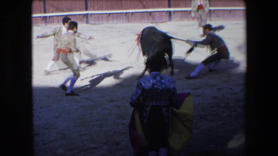 Several young matadors or assistants, including one holding a cape, move around a black bull in the sandy arena during the supplied 1971 Barcelona scene.