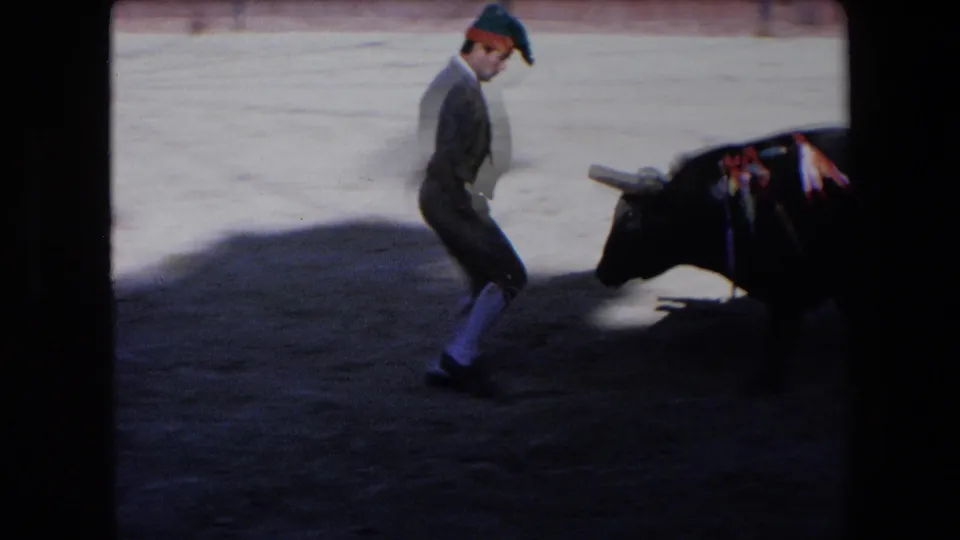 A matador in bullfighting attire runs backward across the arena while a black bull with lowered head advances from the right.