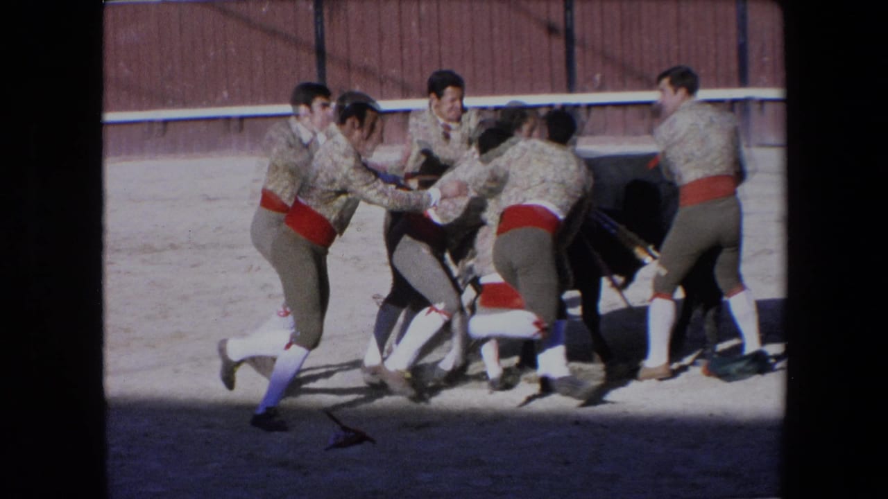 In 1971 Barcelona footage, several bullfighters in beige jackets and red sashes crowd around a black bull near a wooden arena fence.