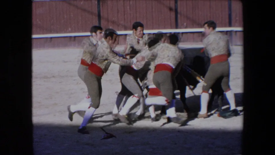 In 1971 Barcelona footage, several bullfighters in beige jackets and red sashes crowd around a black bull near a wooden arena fence.
