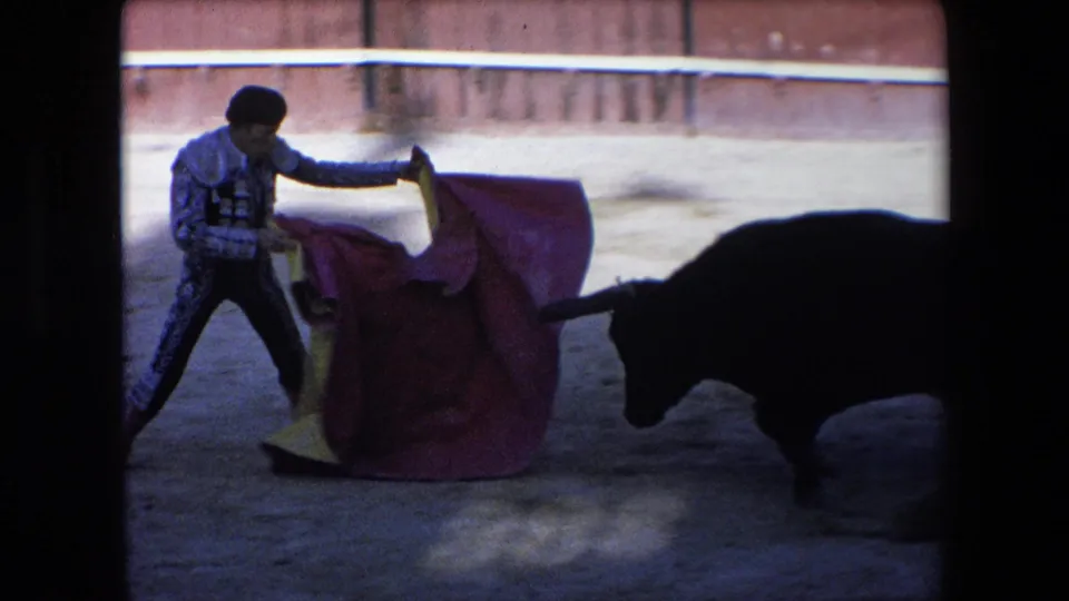 The bullfighter braces with a red and yellow cape as the black horned bull lowers its head close to the cloth in the 1971 Barcelona bullfighting footage.