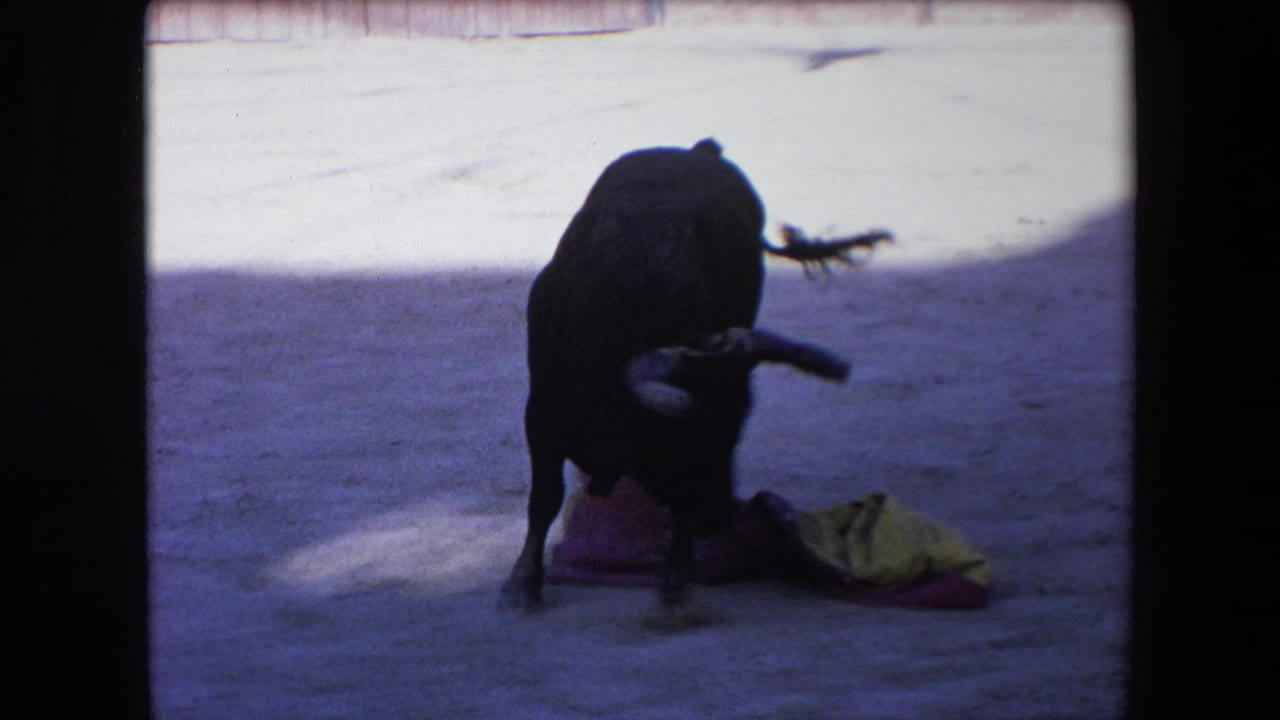 1971 Barcelona 8mm frame showing a black bull standing over or striking a colorful cape on the sand inside a bright bullring.
