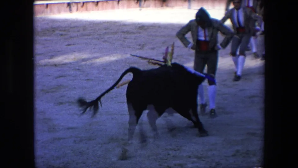 A dark bull charges through the sandy Barcelona bullring with horns extended, passing near a matador in traditional costume while another performer stands nearby.