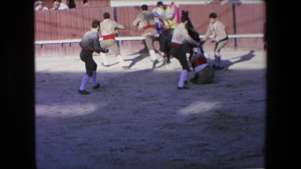 Several bullfighters in an arena run away from a black bull, with one figure down or crouched on the sand near the wooden barrier in the 1971 Barcelona clip.
