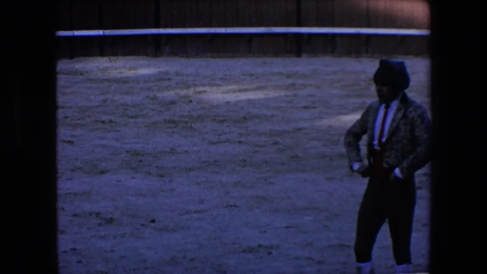 A bullfighter wearing a dark jacket, red sash, white stockings, and a dark head covering stands on the sandy arena floor beneath the shadowed edge of the 8mm frame.