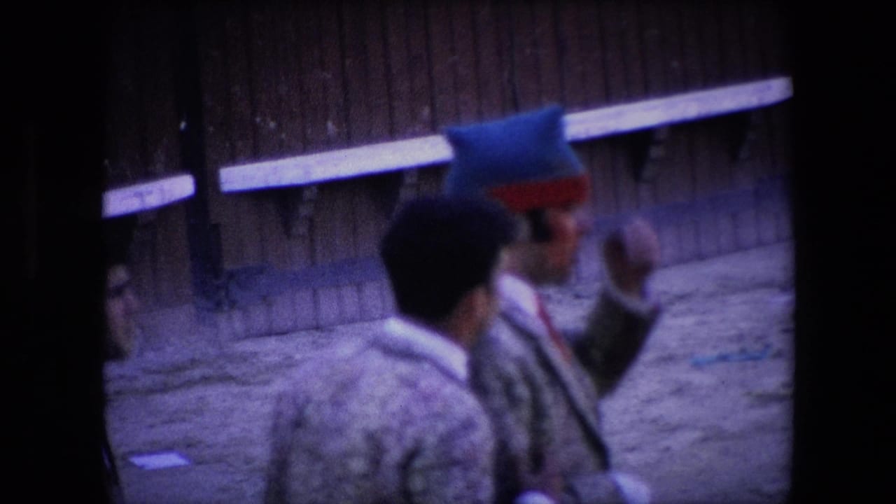 Two men in patterned jackets, one wearing a blue-and-red hat, stand in the sandy bullring area near a wooden wall in the supplied Barcelona 1971 clip.