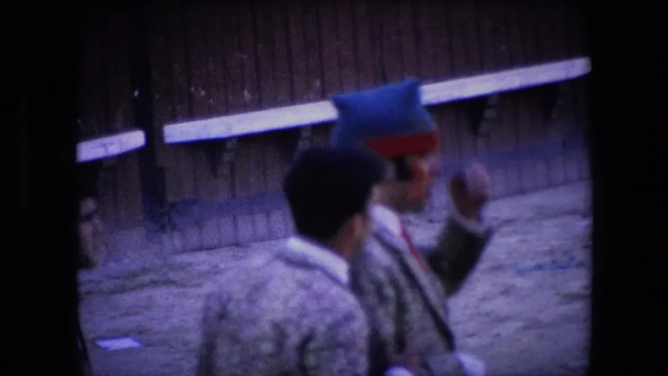 Two men in patterned jackets, one wearing a blue-and-red hat, stand in the sandy bullring area near a wooden wall in the supplied Barcelona 1971 clip.