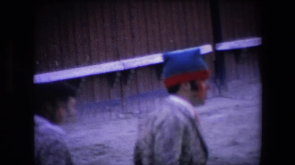 A handheld 8mm frame shows blurred men in patterned bullring clothing moving along the sandy enclosure beside a wooden wall.