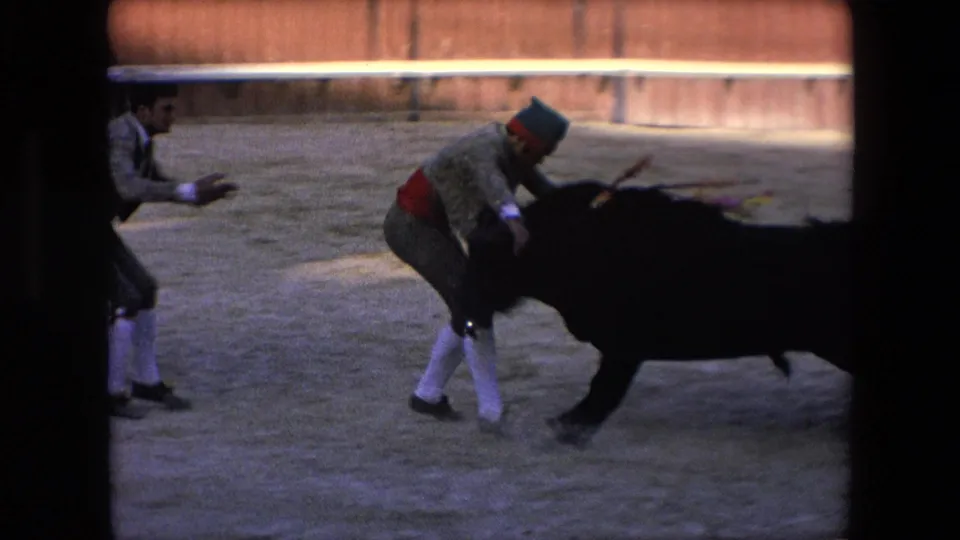 The black bull reaches the matador in the sandy bullring while another arena figure stands nearby, captured in soft 8mm motion blur.