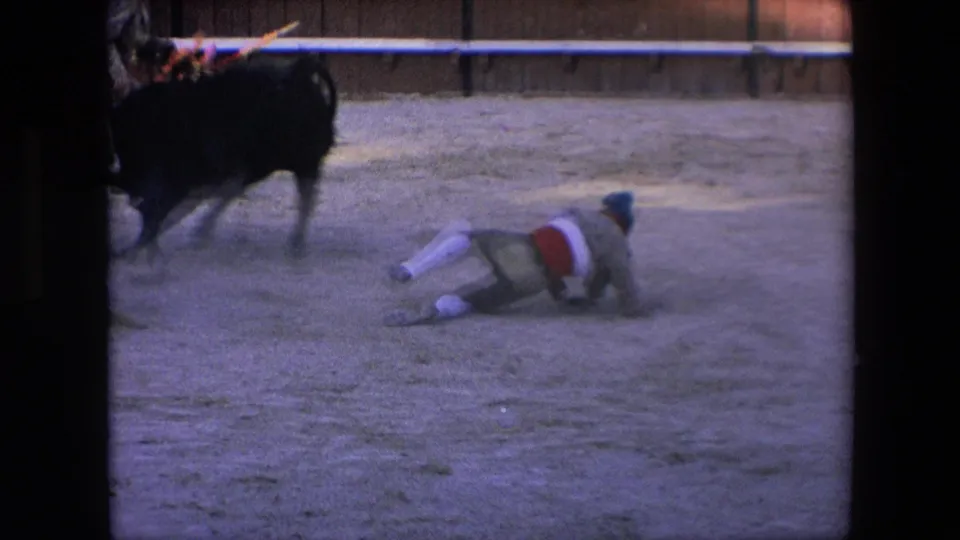 After the collision, the matador lies on the sandy arena floor while the black bull moves away, with the wooden barrier visible in the background.