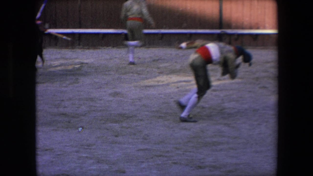 In the supplied 1971 Barcelona bullfighting footage, a costumed performer bends forward in the sand ring while another figure moves along the wooden arena barrier.