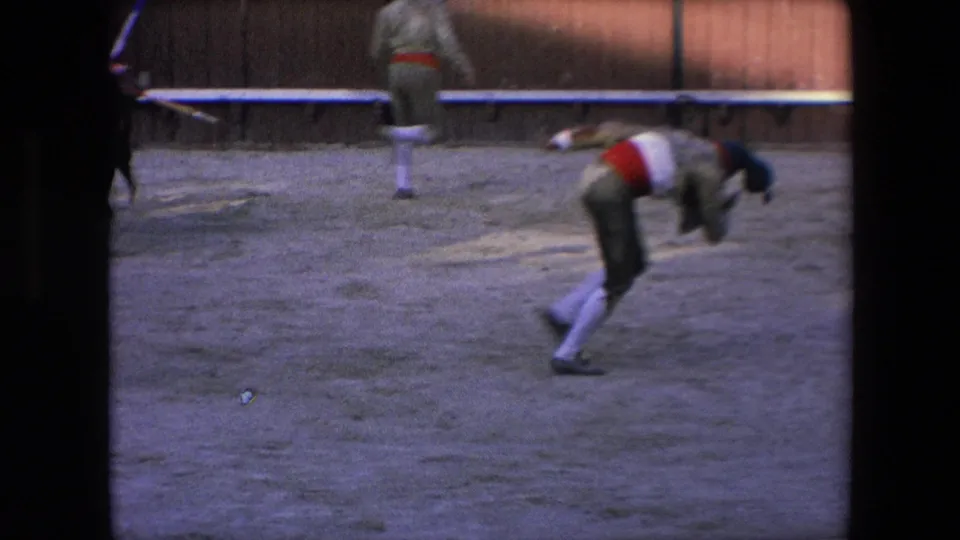 In the supplied 1971 Barcelona bullfighting footage, a costumed performer bends forward in the sand ring while another figure moves along the wooden arena barrier.